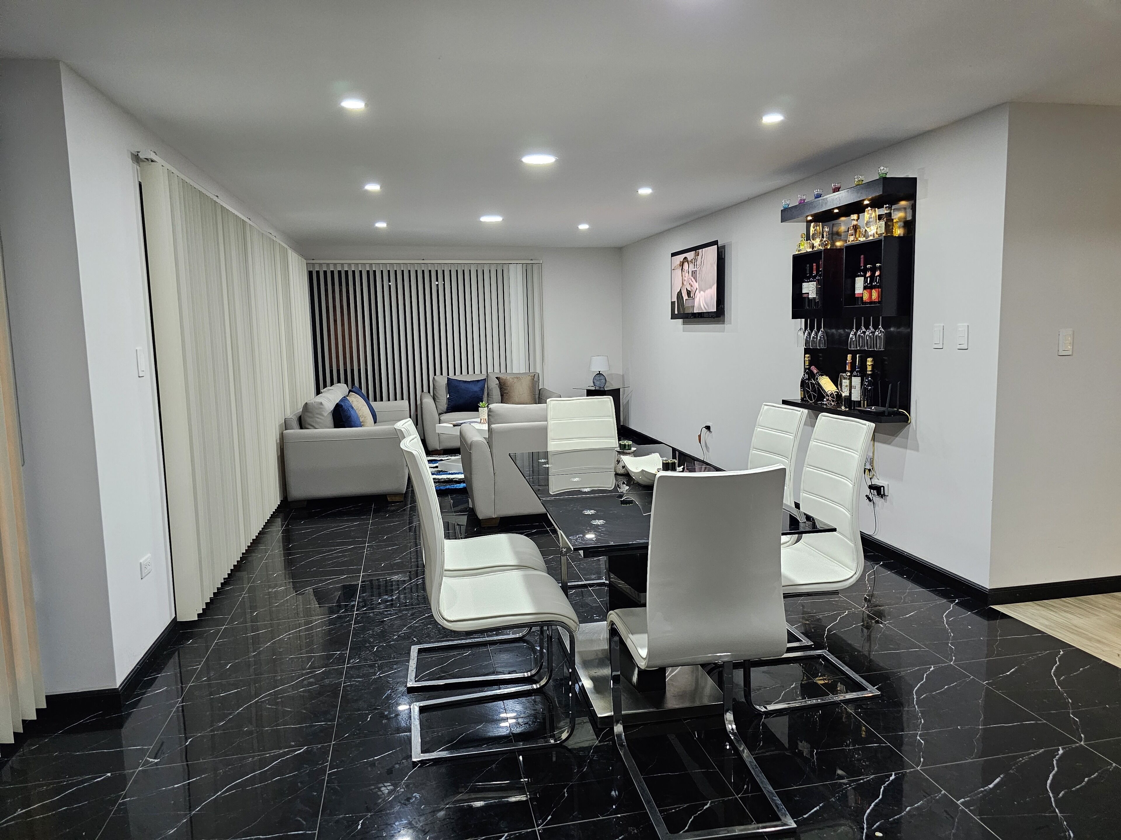 Un comedor moderno con piso de mármol negro, sillas blancas y una mesa larga. Hay una zona de bar con botellas y vasos en la pared.