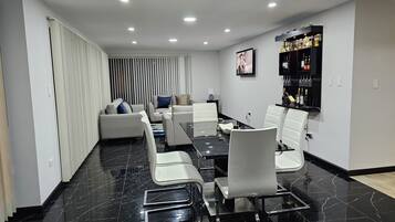 Un comedor moderno con piso de mármol negro, sillas blancas y una mesa larga. Hay una zona de bar con botellas y vasos en la pared.