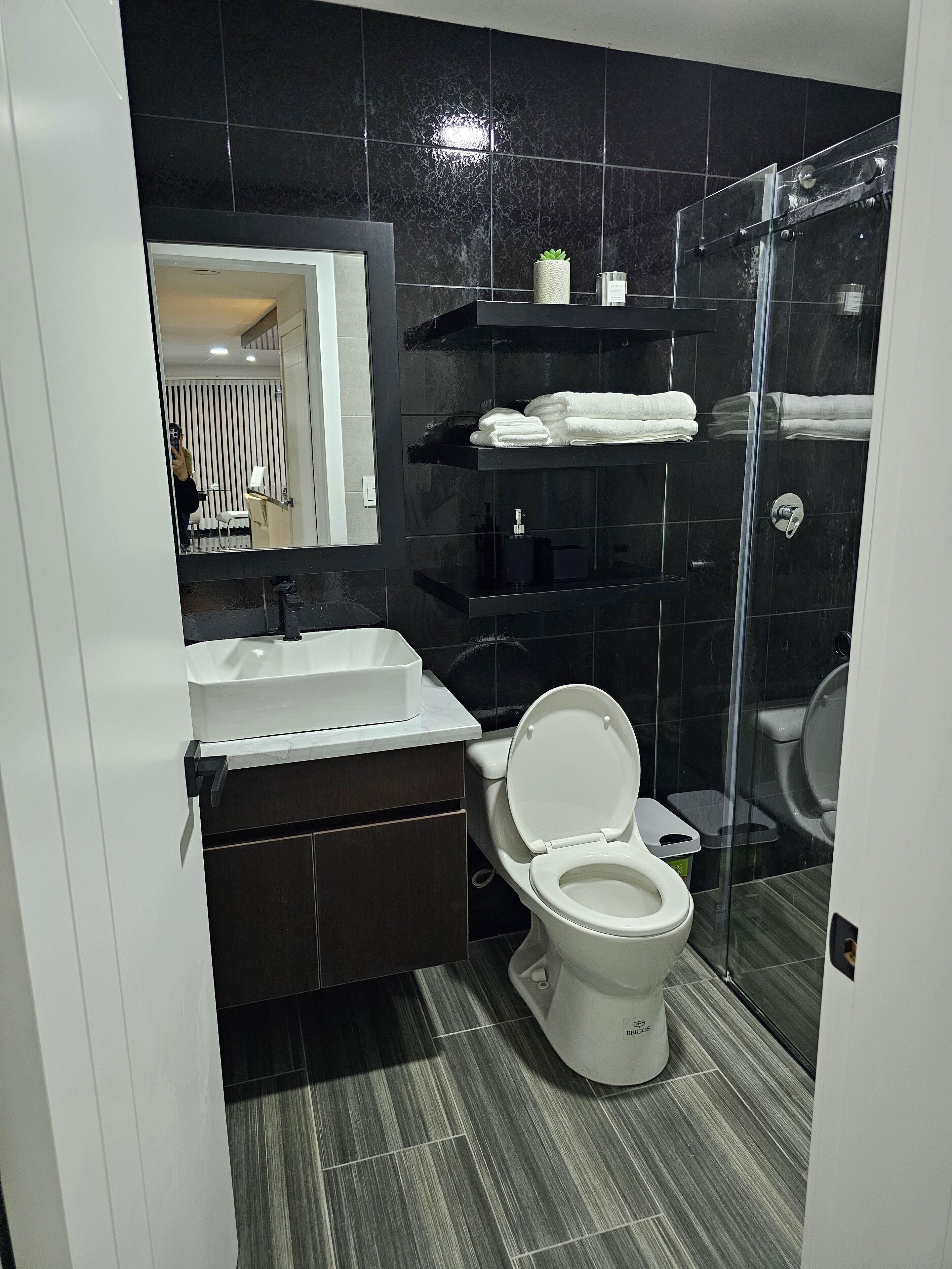 Et moderne badeværelse med en hvid vask, et toilet, en brusekabine i glas og et spejl der reflekterer en stribet væg og en dør.