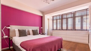 Quarto com cama grande, parede rosa e janela com persianas.