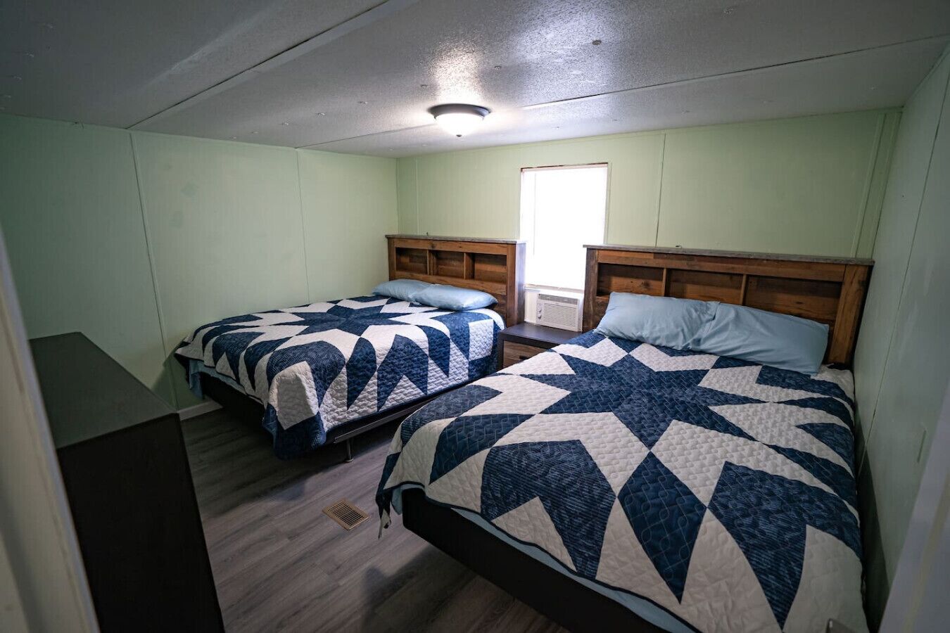 A room with two beds, each with a star-patterned quilt, wooden headboards, and a window providing natural light.