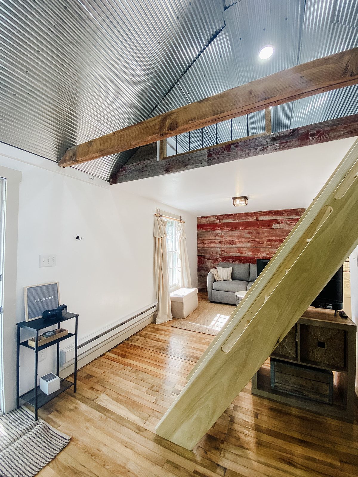 A loft-style room with a wooden floor, a red brick wall, a grey sofa, and a wooden staircase.