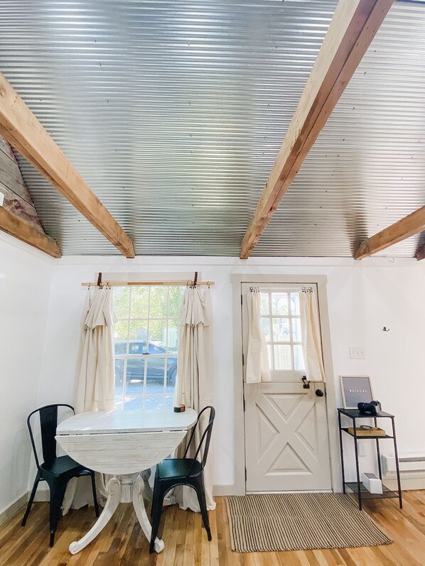 Una habitación con techo de madera, una puerta blanca, una mesa con dos sillas y una mesita auxiliar con un cartel.