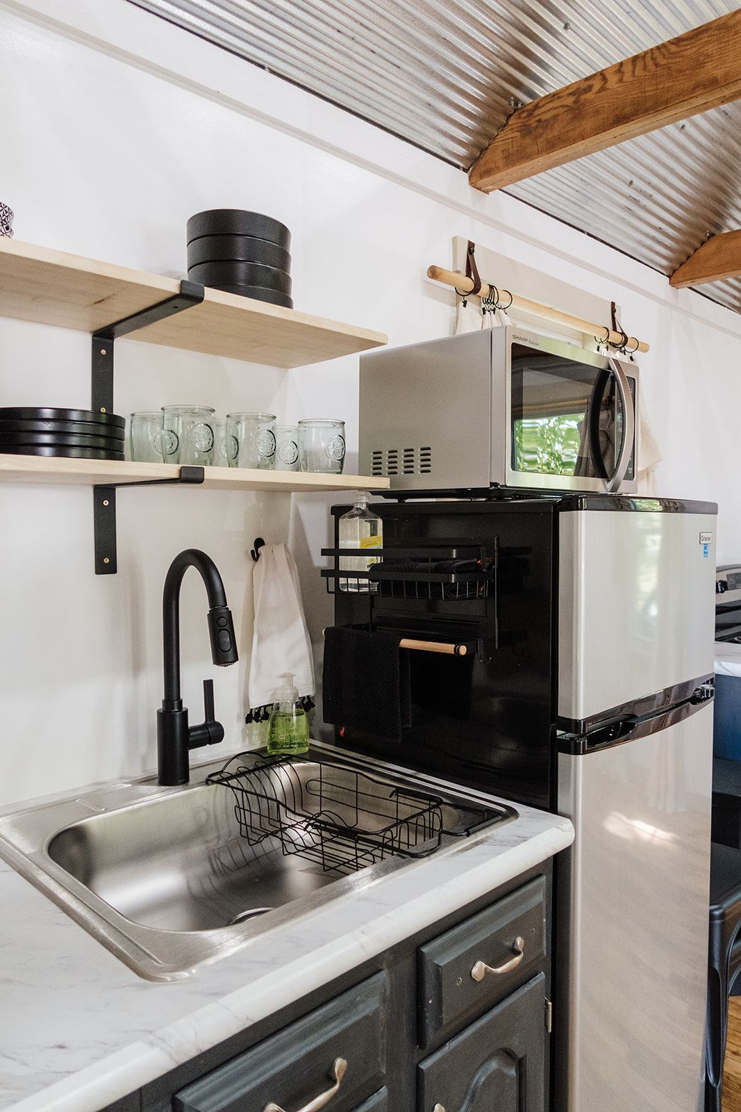 Een kleine keuken met een spoelbak, een magnetron, een inbouwoven en een plank met drinkglazen.