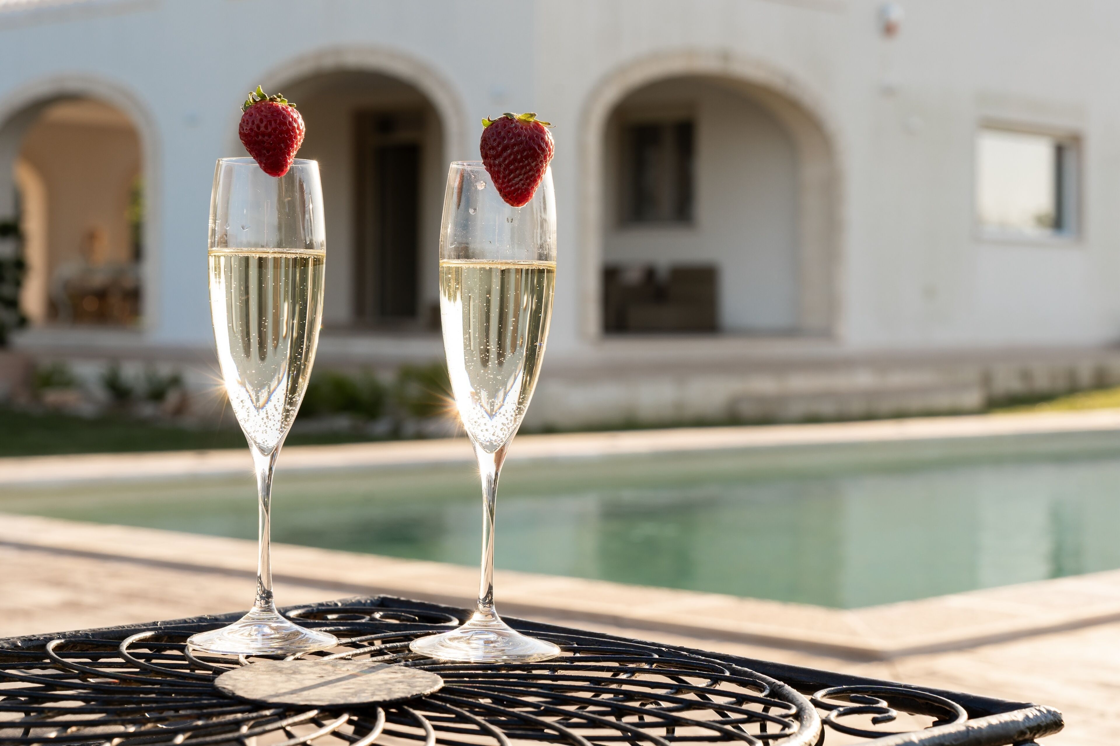 Dos copas de champán con fresas sobre una mesa de metal junto a una piscina, con un edificio al fondo.