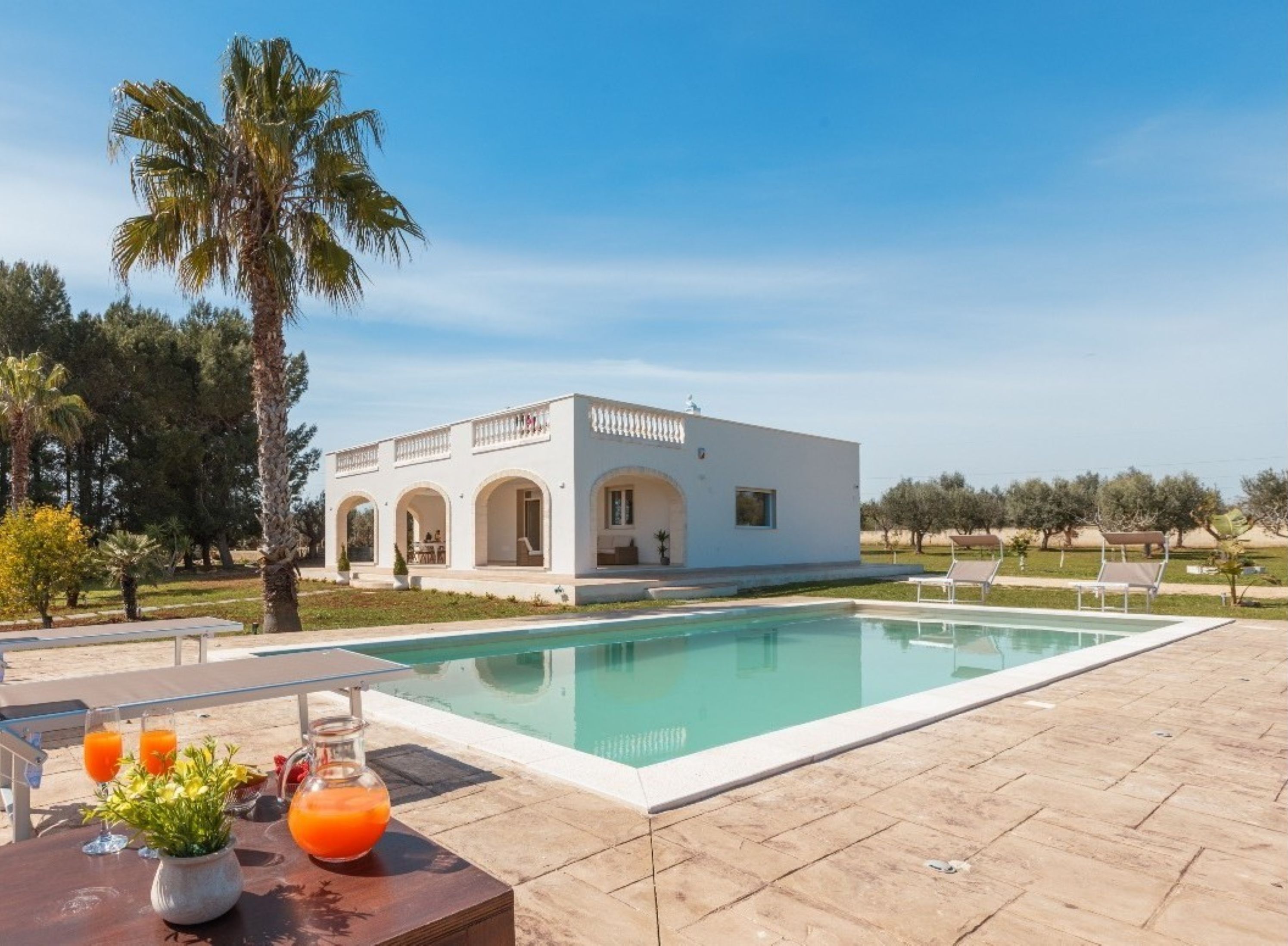 A white villa with a pool, surrounded by palm trees and a clear sky.