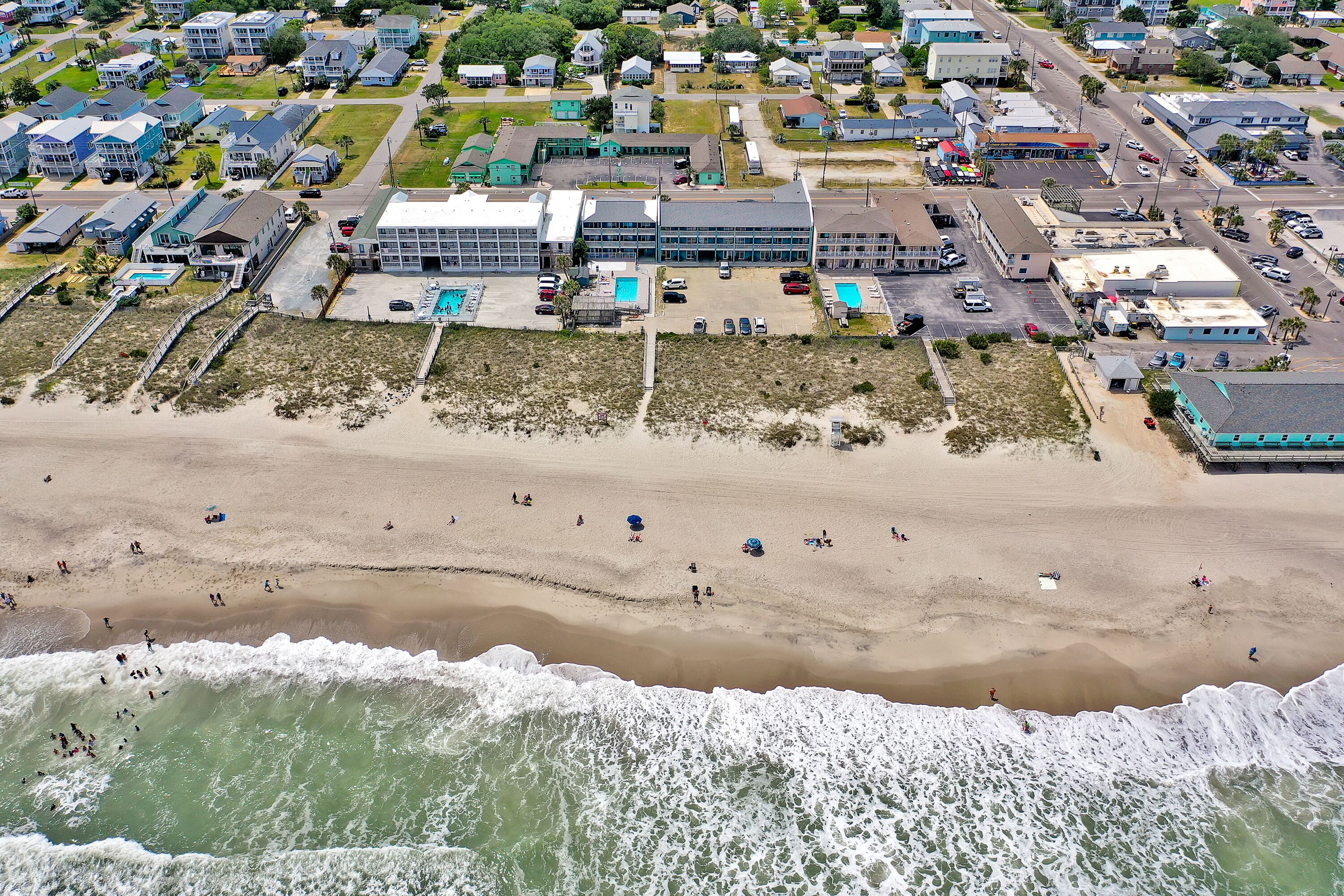 An aerial view of a coastal area with a beach, a large building with multiple floors, and several smaller structures.
