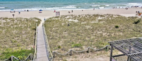 A beach with a wooden boardwalk leading to a wooden structure, surrounded by dunes and a clear sky.