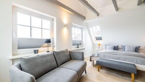 A modern, well-lit room with a grey sofa, a bed with a grey duvet, and a skylight above. - Sylt Seascape IV - HoogeBay - High-quality luxury house for 6 people with sauna, (Sylt)