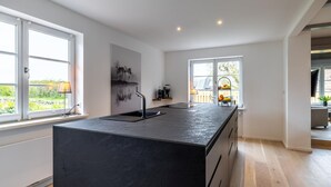 A modern kitchen with a large island, two windows, and a view of the outdoors. - Sylt Seascape IV - HoogeBay - High-quality luxury house for 6 people with sauna, (Sylt)