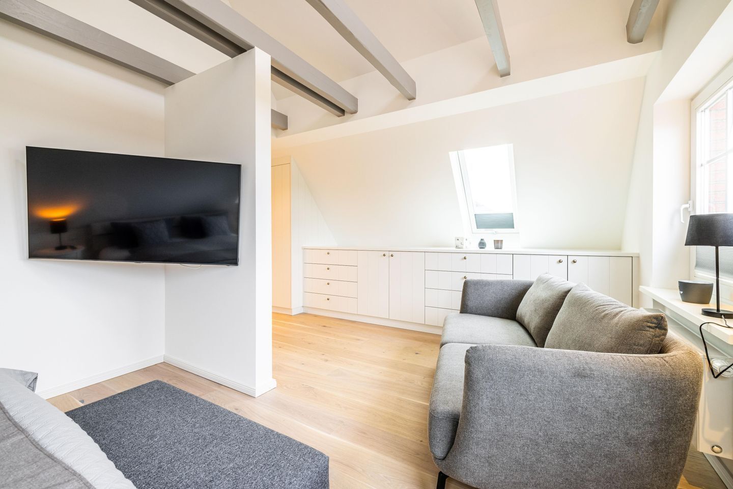 Un salon moderne avec un téléviseur à écran plat fixé au mur, un canapé gris, un petit tapis et un coin cuisine à l’arrière-plan.