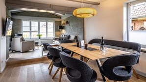 A modern dining area with a wooden table and black chairs, a hanging light fixture, and a view of the kitchen and living space. - Sylt Seascape IV - HoogeBay - High-quality luxury house for 6 people with sauna, (Sylt)