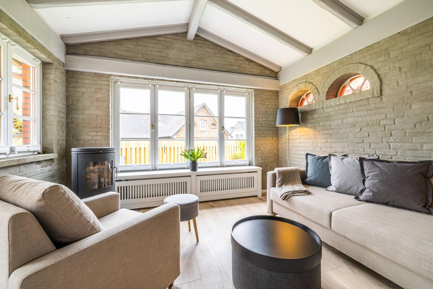 A cozy living room with a high ceiling, exposed brick walls, a large window, and a fireplace.
