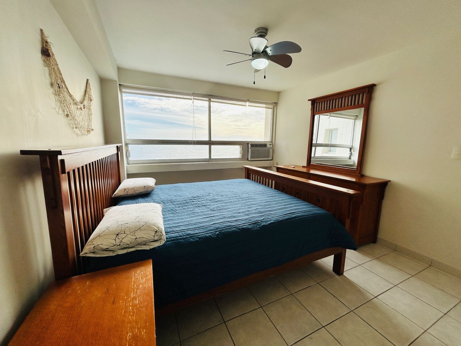 A bedroom with a bed, a wooden headboard, a bench, a ceiling fan, a window with a view, and a mirror.