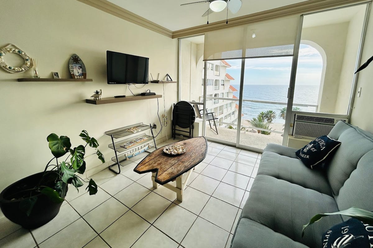 A living room with a sofa, a coffee table, a TV, and a balcony with a view of the sea.