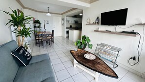 Una sala de estar moderna con un sofá, una mesa de centro y plantas. Se observa un área de cocina al fondo, con un televisor montado en la pared.