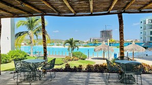 A covered outdoor seating area with a pool view, featuring palm trees and a clear sky. - Blue Lagoon (Mazatlán)