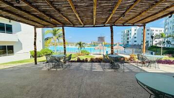 A covered outdoor area with tables and chairs, overlooking a pool and a beach.