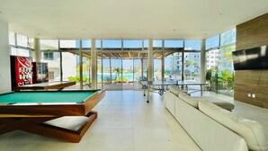 A modern living room with a pool table, a sofa, and a large window offering a view of a pool and buildings. - Blue Lagoon (Mazatlán)