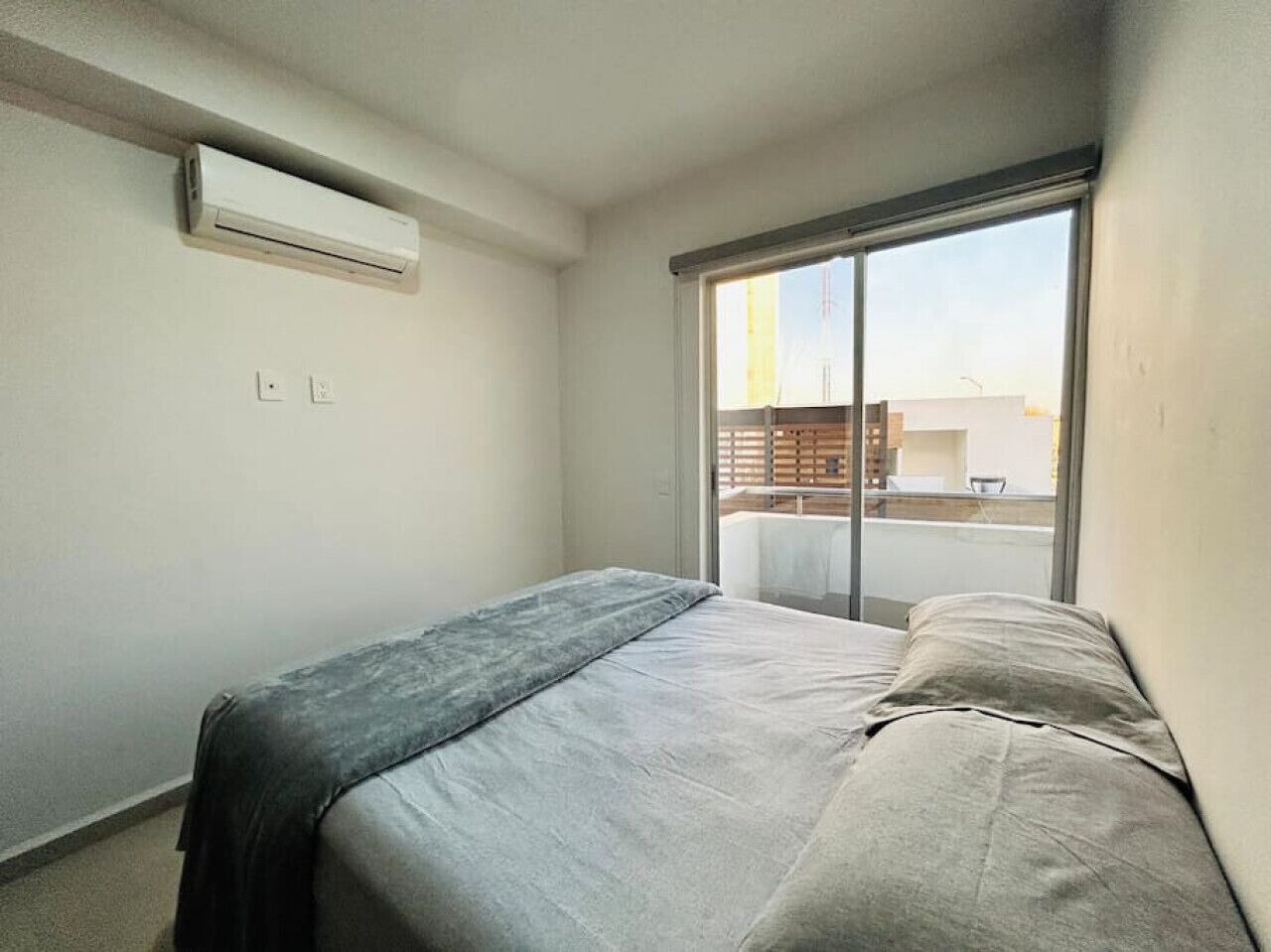 Un dormitorio con cama, aire acondicionado y un ventanal con vistas a edificios.