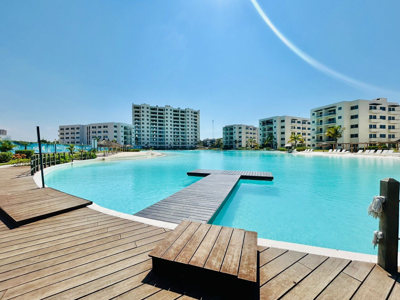 A large, clear swimming pool with wooden deck areas, surrounded by modern buildings.