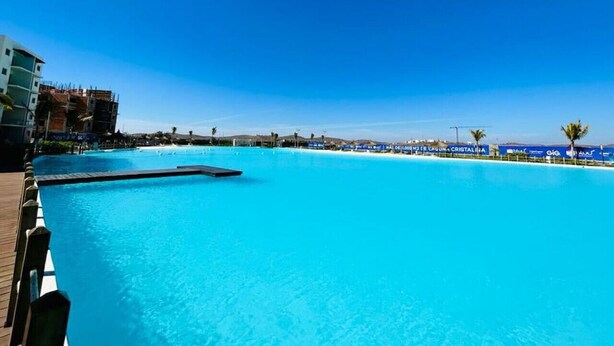 A large, clear blue pool with a wooden deck on one side and a beach area with umbrellas in the background. - Blue Lagoon (Mazatlán)