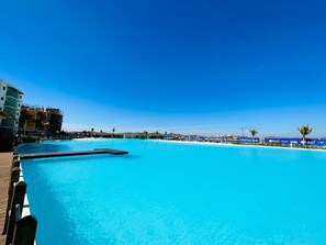 A large, clear blue pool with a wooden deck on one side and a beach area with umbrellas in the background.