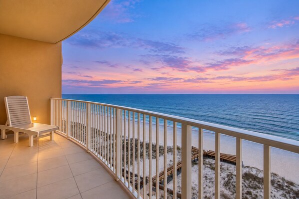 A balcony with a white chair, a railing, and a view of the ocean at sunset.
