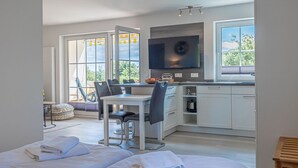 A modern, open-plan bedroom and kitchen with a large window, a dining area, and a flat-screen TV. - fewo1846 - South view (App. 689) / Studio apartment with balcony on the mezzanine floor - (Harrislee)