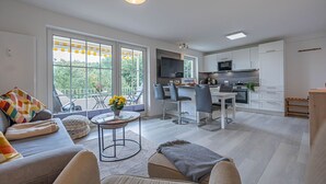 A modern living room with a sofa, a coffee table, and a dining area with chairs. There is a kitchen with cabinets and appliances in the background. - fewo1846 - South view (App. 689) / Studio apartment with balcony on the mezzanine floor - (Harrislee)