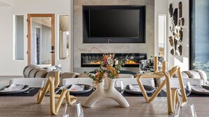 Een moderne woonkamer met een grote flatscreen televisie aan de muur, een open haard en een eetgedeelte met bijzondere stoelen.