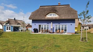 Ein Haus mit einem Strohdach, blauer Fassade und einer Terrasse mit Gartenmöbeln.