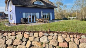 Ein Haus mit Strohdach, einer Steinsäule im Vordergrund und einer Terrasse mit Gartenmöbeln.