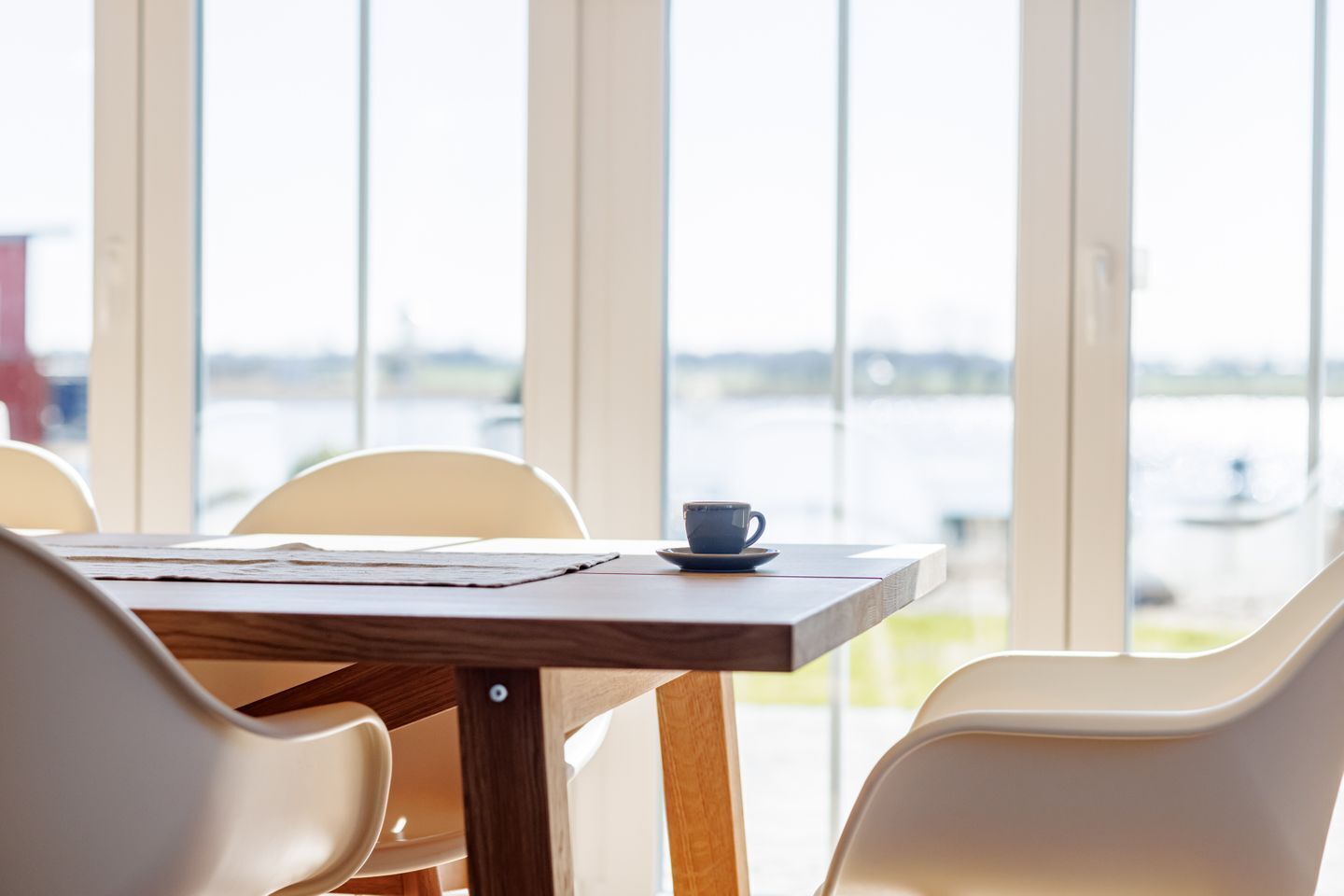 A table with a cup and saucer, chairs, and a view of a body of water through large windows.