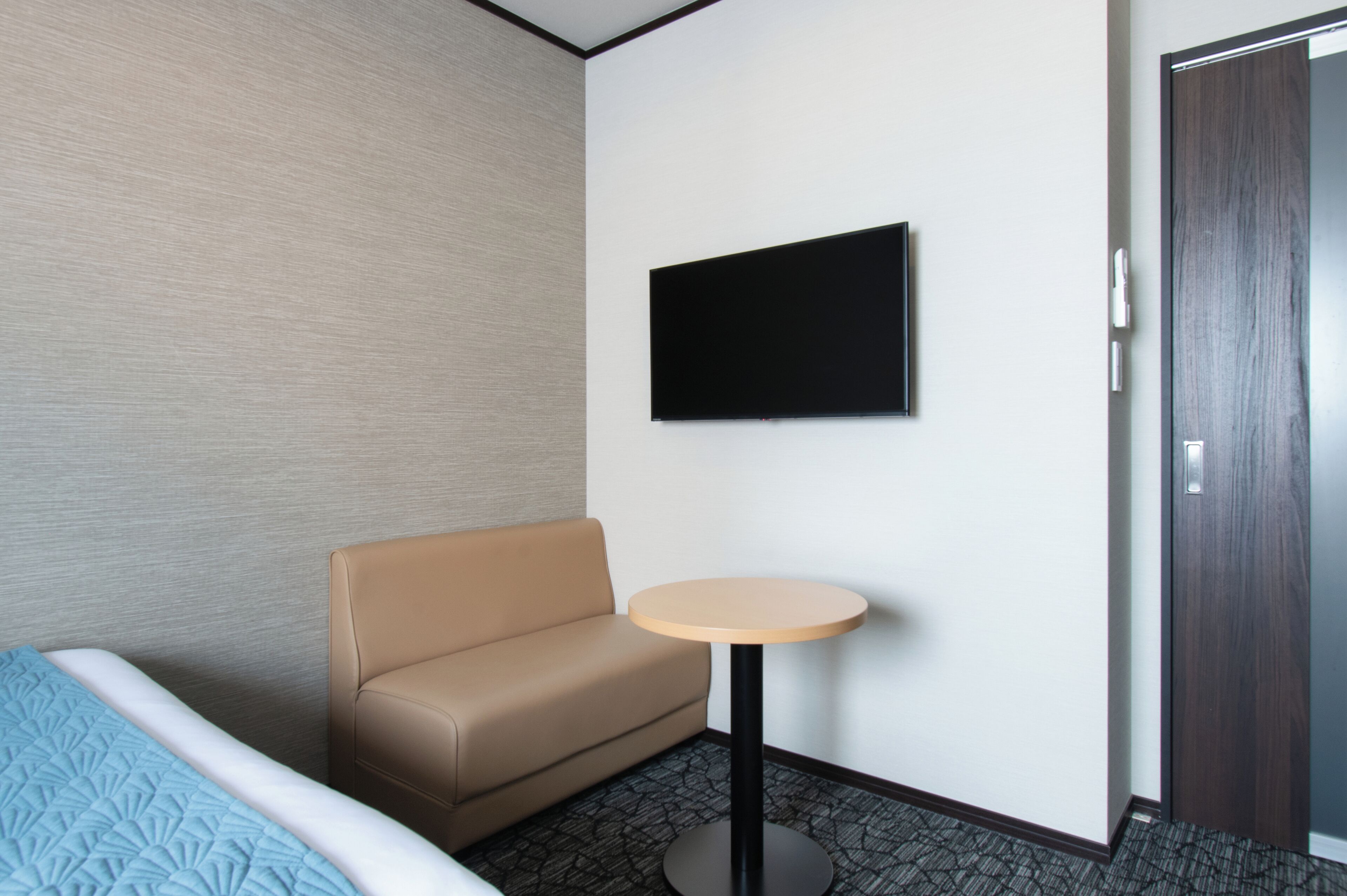 A hotel room with a bed, a sofa, a small round table, a flat-screen TV mounted on the wall, and a door.