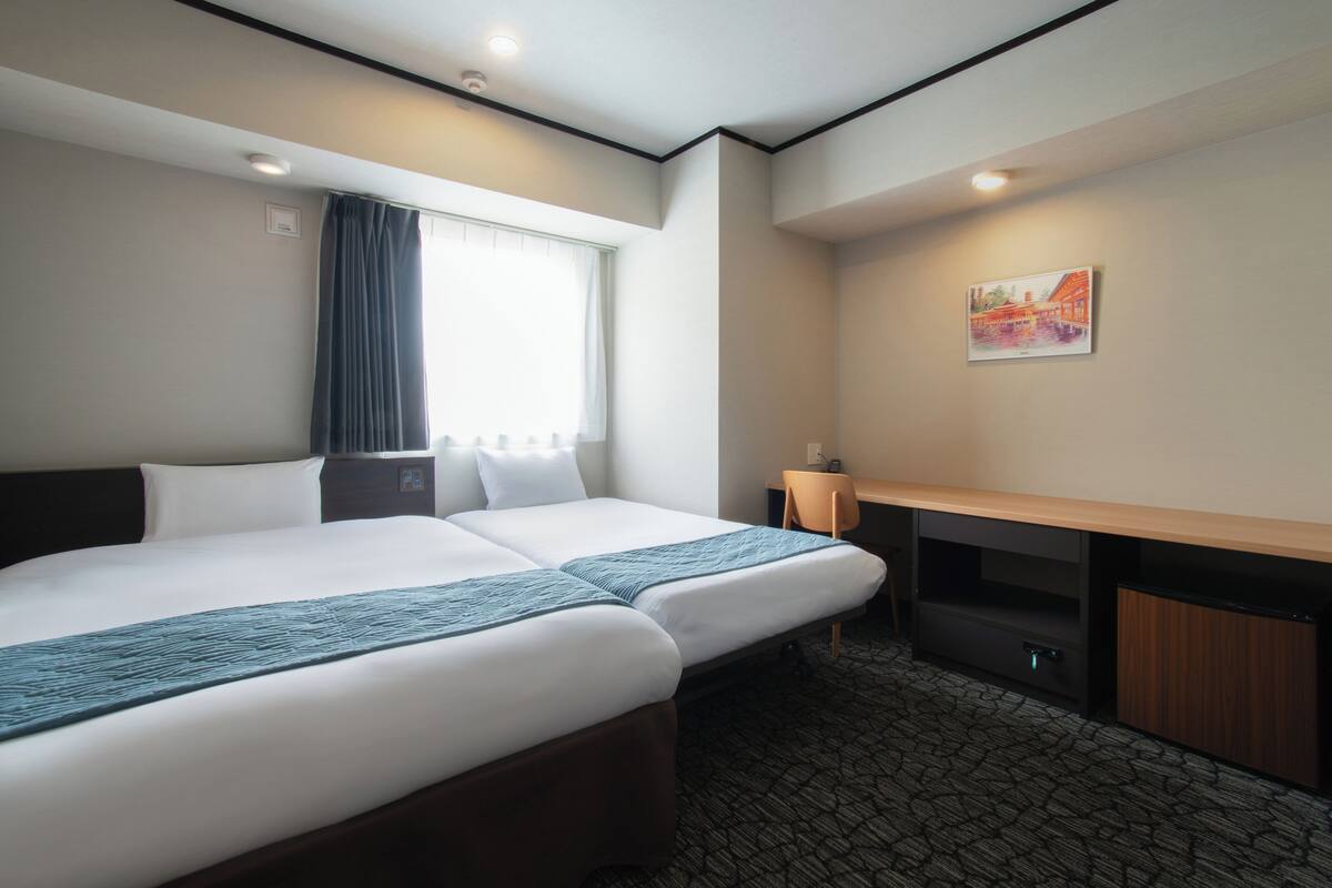 A hotel room with two beds, a desk, a chair, a small cabinet, and a painting on the wall.