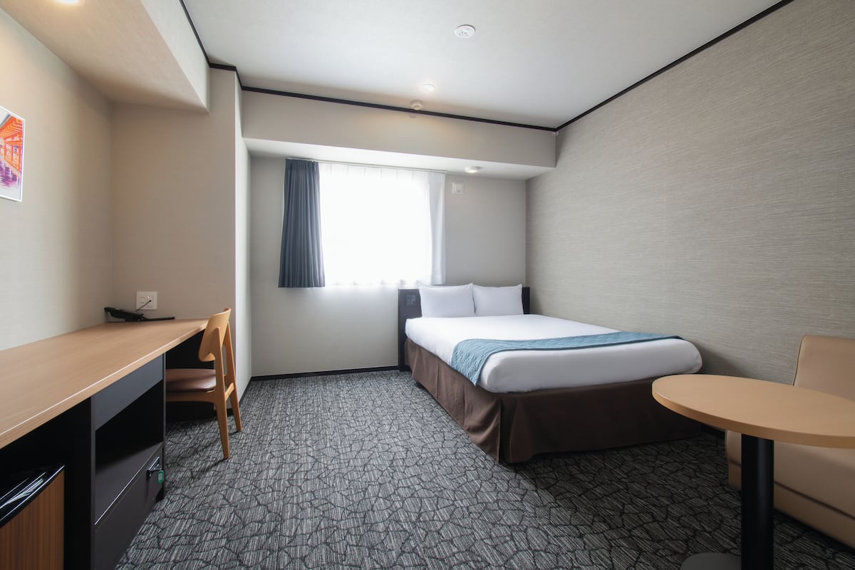A hotel room with a bed, a desk, a chair, a small table, and a window with curtains.