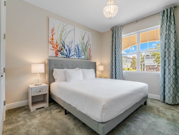 A neatly made bed in a hotel room with a large window, a decorative painting above the bed, and a chandelier.