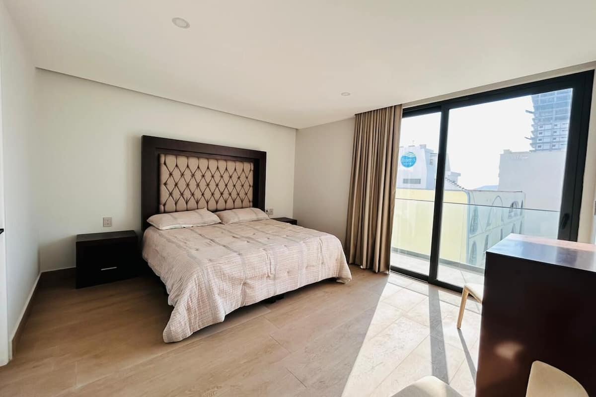 Un dormitorio con cama, mesita de noche, escritorio y un ventanal con vistas a edificios.