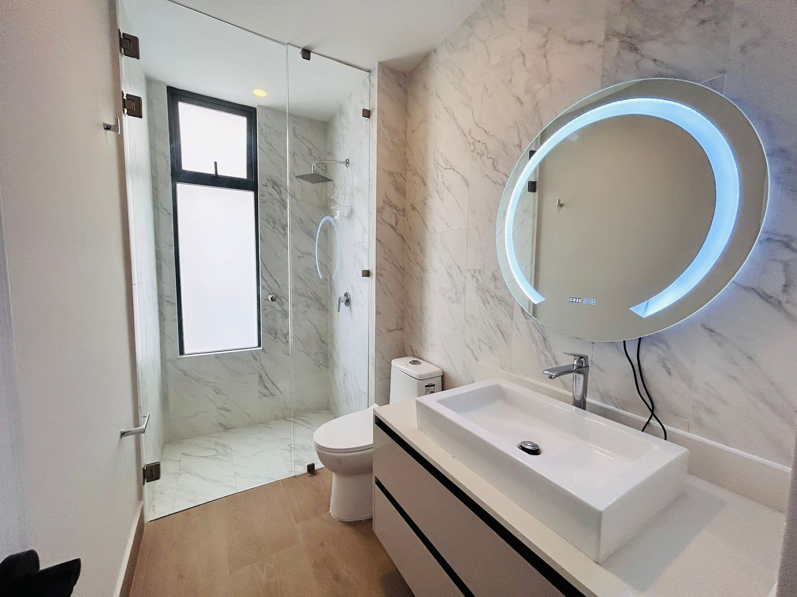 Un baño moderno con ducha envuelta en vidrio, inodoro blanco, lavamanos blanco con encimera de mármol y un espejo redondo con luz.