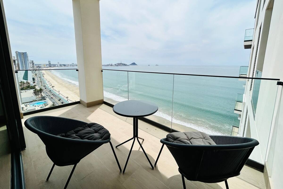 Balcón con dos sillas negras y una mesa pequeña, con vista a una playa y al océano.