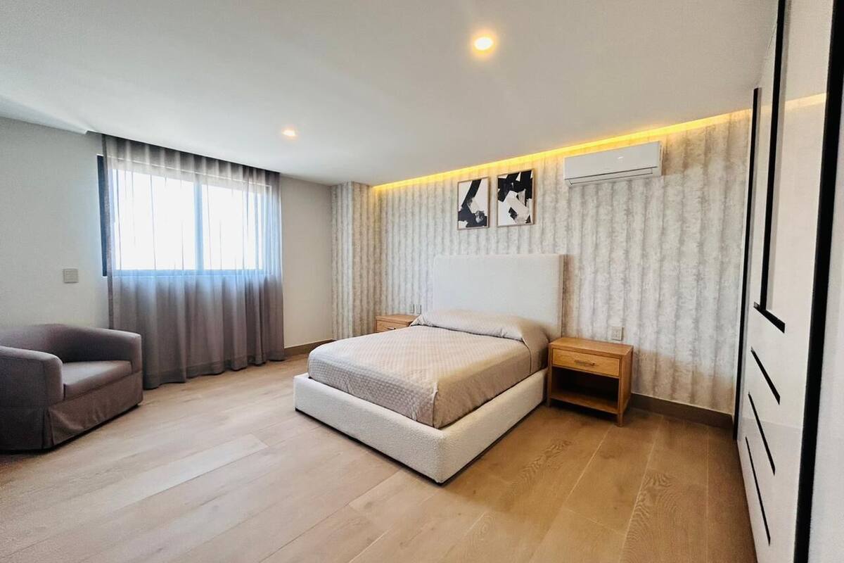 Un dormitorio moderno con una cama grande, una mesita de noche, una silla y un ventanal con cortinas.