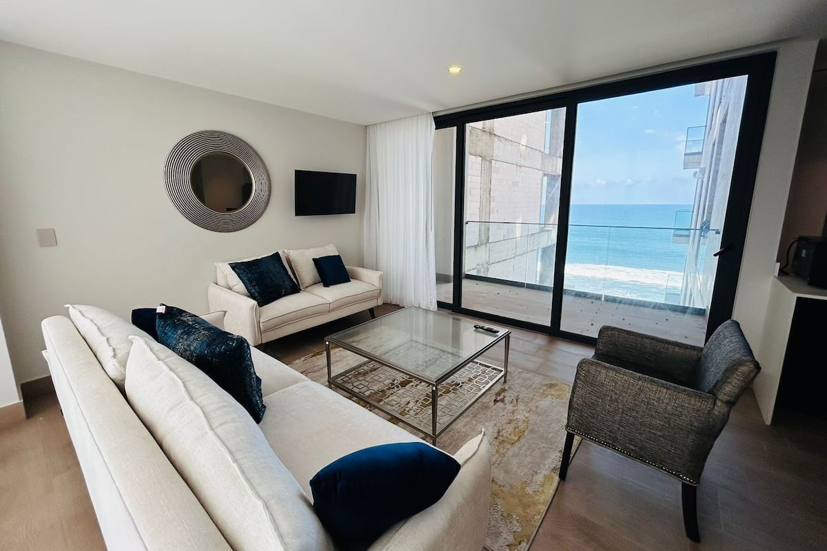 Una sala moderna con sofá, sillón y mesa de centro, un ventanal con vista al océano y un televisor de pantalla plana en la pared.