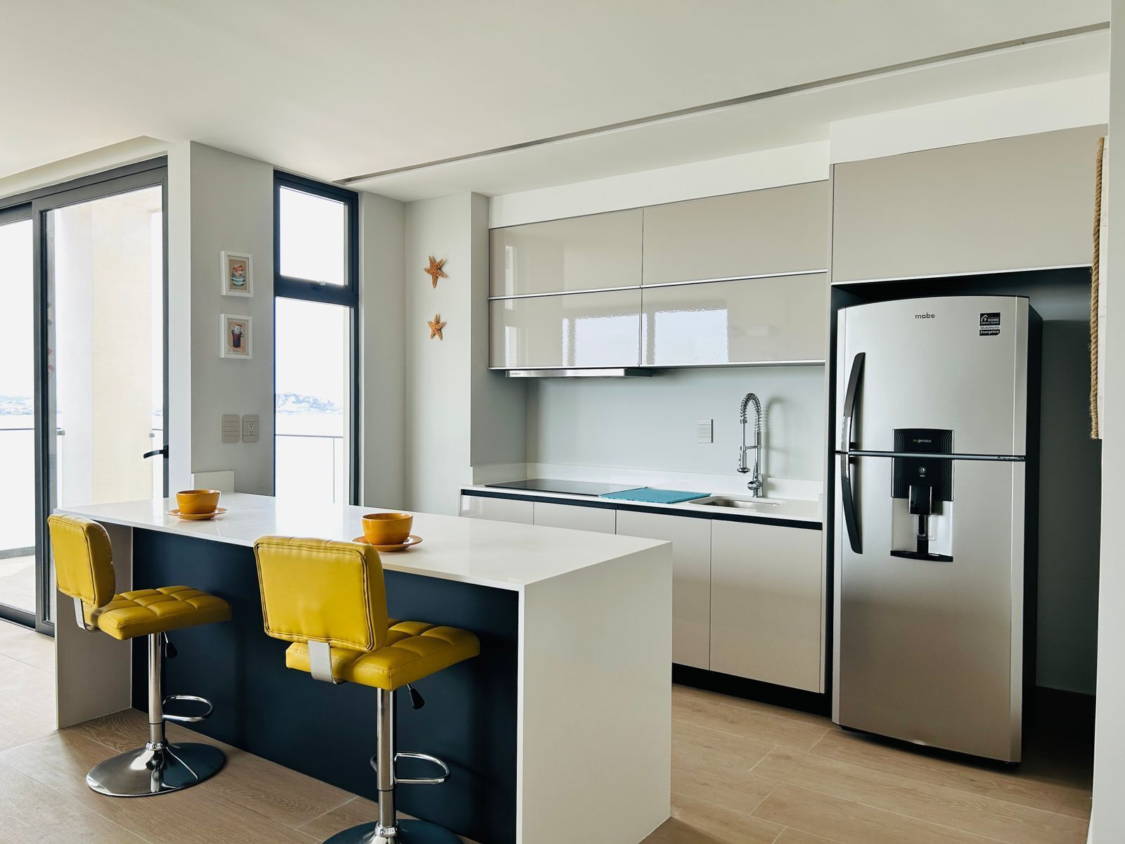 A modern kitchen with a central island, bar stools, a refrigerator, and a window providing natural light.