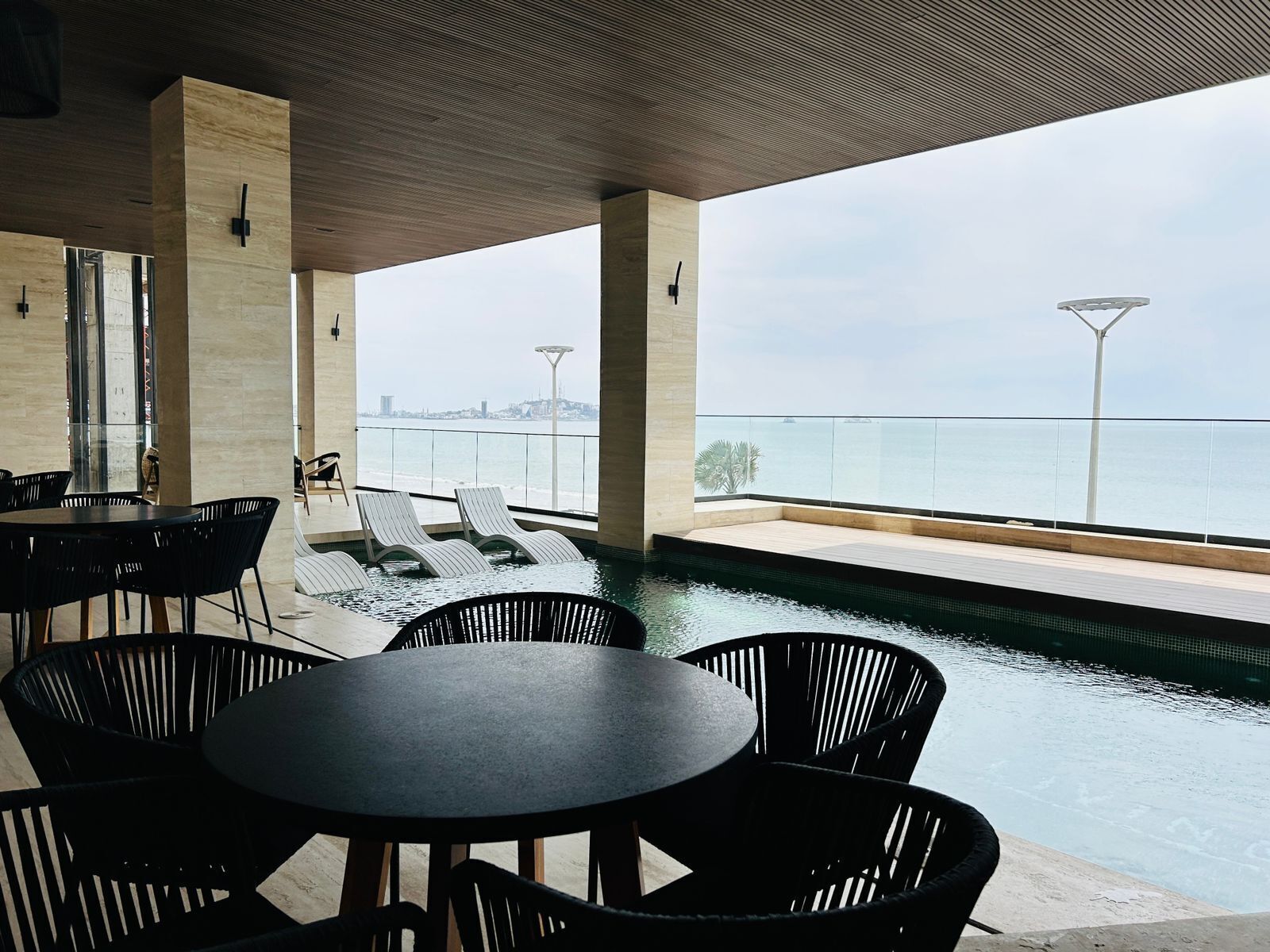 A modern outdoor pool area with lounge chairs and tables, a view of the sea, and a cityscape in the background.