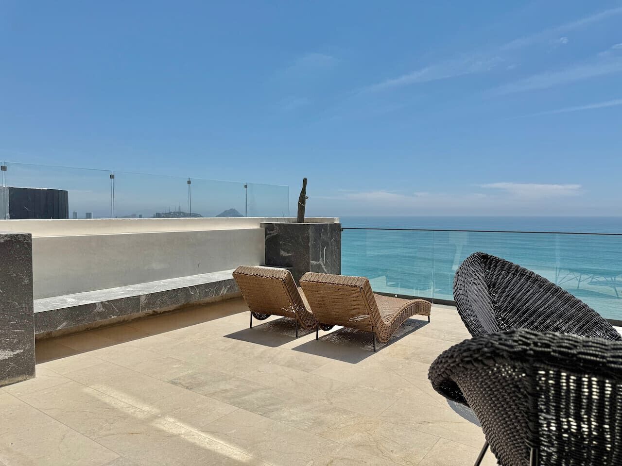 Eine Dachterrasse mit zwei Korbsessel-Loungemöbeln, die auf eine Glasbrüstung mit Blick aufs Meer ausgerichtet sind.