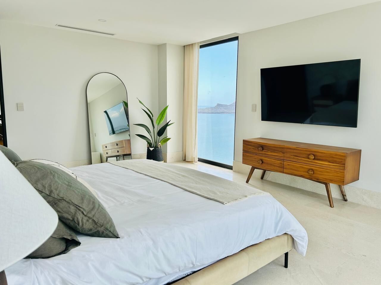 A modern bedroom with a large bed, a wooden dresser, a flat-screen TV, a large mirror, and a view of the sea.