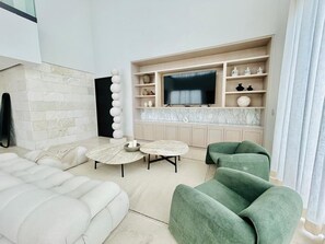 Un salon moderne comprenant un canapé, deux tables rondes, un téléviseur à écran plat et un coin cuisine doté de plans de travail en marbre.