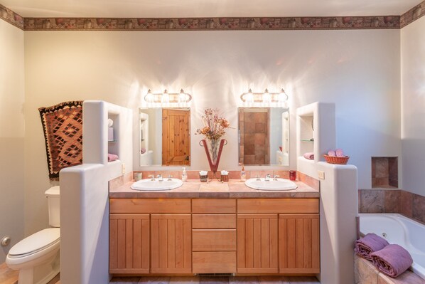 A bathroom with two sinks, a wooden vanity, a mirror with lights, a toilet, and a bathtub.
