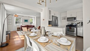 Une cuisine moderne équipée d’appareils électroménagers en acier inoxydable, un coin repas avec une longue table pouvant accueillir six personnes, et un salon confortable doté d’une cheminée.